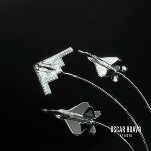 May include: Three silver-colored model fighter jets against a black background. The jets are attached to thin, curved metal rods. The jets include a B-2 bomber and two F-22 Raptors. The image includes the text "OSCAR BRAVO STUDIO".