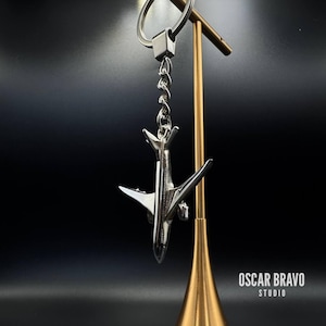 May include: A silver airplane keychain with a detailed design, hanging from a metal ring and chain. The keychain is suspended against a dark background, with the text "OSCAR BRAVO STUDIO" visible.