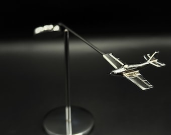 May include: A silver-colored model airplane mounted on a slender, curved metal stand. The airplane has detailed wings and tail, with a polished finish. The stand features a circular base and a curved arm, set against a dark background. The text "OSCAR BRAVO STUDIO" is visible.