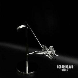 Könnte beinhalten: Ein silbernes Modell eines Kampfjets auf einem silbernen Ständer. Der Jet schwebt in der Luft, wobei der Ständer ihn hält. Der Hintergrund ist schwarz.