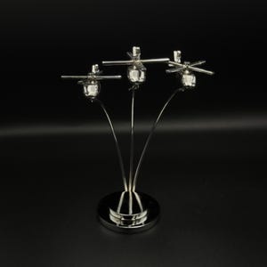 May include: A silver-colored metal sculpture featuring three miniature helicopter models. Each helicopter has a detailed body and rotor blades, supported by curved metal stems rising from a circular base. The sculpture is set against a black background.