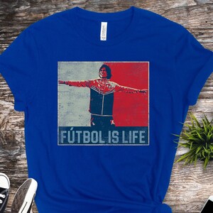Pu&ograve; includere: T-shirt blu reale con grafica retr&ograve; di un calciatore con le braccia tese, su sfondo rosso e blu. Il testo "F&Uacute;TBOL IS LIFE" &egrave; stampato sotto. Sono visibili scarpe da ginnastica nere e uno smartphone.