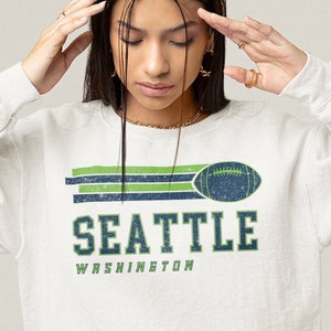 May include: A white sweatshirt with a green and blue graphic print of a football and the text "SEATTLE WASHINGTON".