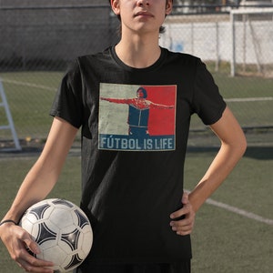 Pu&ograve; includere: Maglietta nera con grafica di un calciatore con le braccia tese, su sfondo rosso e blu. Il testo "F&Uacute;TBOL IS LIFE" &egrave; sotto l'immagine. Un pallone da calcio &egrave; tenuto tra le mani.