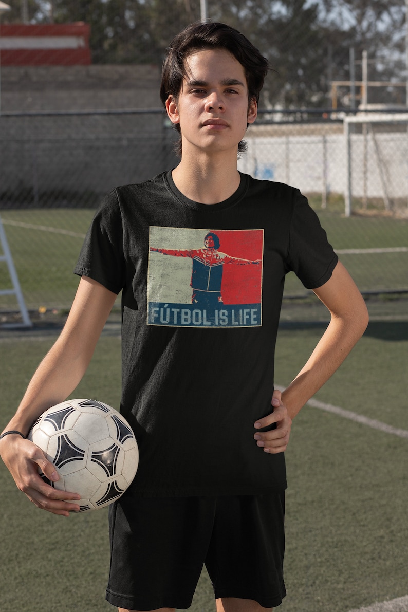 Pu&ograve; includere: Maglietta nera con grafica di un calciatore con le braccia tese, su sfondo rosso e blu. Il testo "F&Uacute;TBOL IS LIFE" &egrave; sotto l'immagine. Un pallone da calcio &egrave; tenuto tra le mani.
