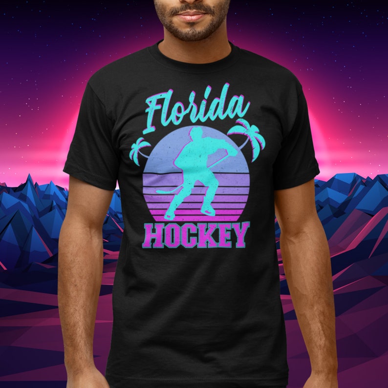 Puede incluir: Camiseta negra con un gr&aacute;fico de estilo retro de un jugador de hockey frente a una puesta de sol con palmeras. El texto "Florida Hockey" est&aacute; impreso debajo del gr&aacute;fico.