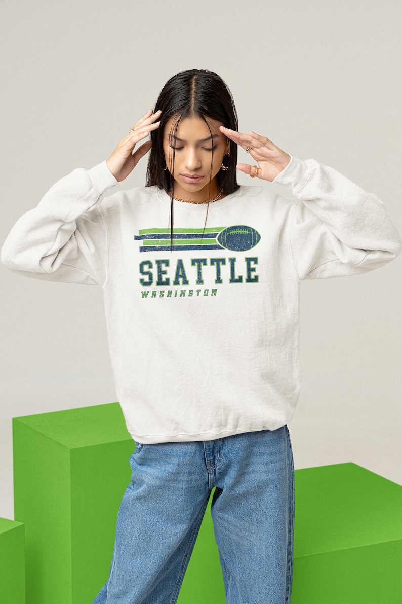 May include: A white sweatshirt with a green and blue graphic print of a football and the text "SEATTLE WASHINGTON".