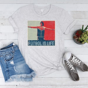 Pu&ograve; includere: T-shirt grigio chiaro con grafica vintage di una persona con le braccia tese, su sfondo rosso e blu. Il testo "F&Uacute;TBOL IS LIFE" &egrave; stampato sotto. La maglietta &egrave; abbinata a pantaloncini di jeans e scarpe da ginnastica grigie.