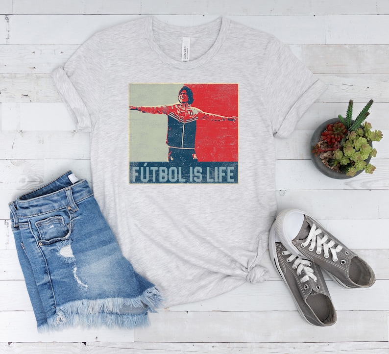 Pu&ograve; includere: T-shirt grigio chiaro con grafica vintage di una persona con le braccia tese, su sfondo rosso e blu. Il testo "F&Uacute;TBOL IS LIFE" &egrave; stampato sotto. La maglietta &egrave; abbinata a pantaloncini di jeans e scarpe da ginnastica grigie.