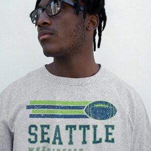 May include: A gray sweatshirt with a vintage-style graphic of a football and the text "SEATTLE WASHINGTON". The graphic is in green, blue, and white.