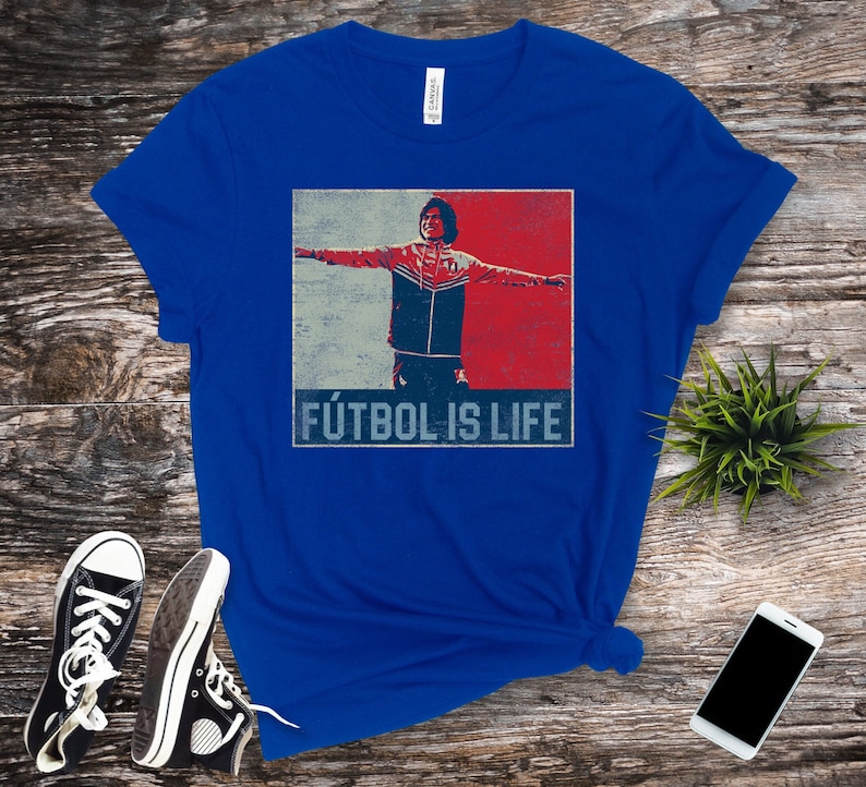 Pu&ograve; includere: T-shirt blu reale con grafica retr&ograve; di un calciatore con le braccia tese, su sfondo rosso e blu. Il testo "F&Uacute;TBOL IS LIFE" &egrave; stampato sotto. Sono visibili scarpe da ginnastica nere e uno smartphone.