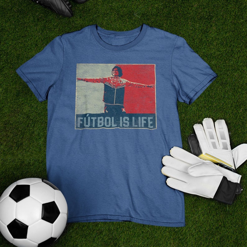 Pu&ograve; includere: Maglietta blu con una grafica di una persona con le braccia tese, che indossa una giacca rossa, bianca e blu. Il testo "F&Uacute;TBOL IS LIFE" &egrave; sotto l'immagine. Un pallone da calcio, scarpe da calcio e un paio di guanti da calcio bianchi e neri sono su una superficie verde.