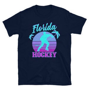 Puede incluir: Una camiseta azul marino con un gr&aacute;fico retro de un jugador de hockey frente a una puesta de sol con palmeras. El texto "Florida Hockey" est&aacute; impreso debajo del gr&aacute;fico.
