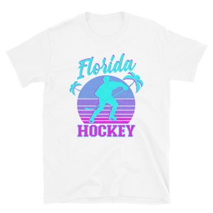 Puede incluir: Camiseta blanca con un gr&aacute;fico azul y rosa de un jugador de hockey frente a una puesta de sol con palmeras y el texto "Florida Hockey".