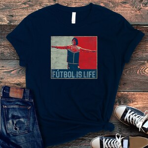Pu&ograve; includere: T-shirt blu navy con una grafica di una persona con le braccia tese, su sfondo rosso, bianco e blu. Il testo "F&Uacute;TBOL IS LIFE" &egrave; stampato sotto. La maglietta &egrave; abbinata a jeans blu e scarpe da ginnastica nere.