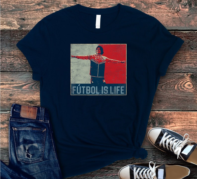Pu&ograve; includere: T-shirt blu navy con una grafica di una persona con le braccia tese, su sfondo rosso, bianco e blu. Il testo "F&Uacute;TBOL IS LIFE" &egrave; stampato sotto. La maglietta &egrave; abbinata a jeans blu e scarpe da ginnastica nere.