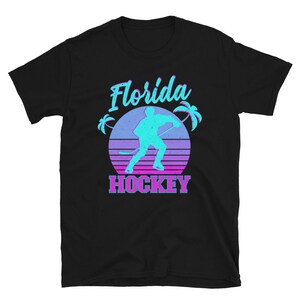 Puede incluir: Camiseta negra con un gr&aacute;fico retro de un jugador de hockey frente a una puesta de sol con palmeras. El texto "Florida Hockey" est&aacute; impreso en rosa y azul.