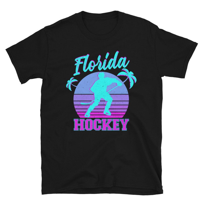 Puede incluir: Camiseta negra con un gr&aacute;fico retro de un jugador de hockey frente a una puesta de sol con palmeras. El texto "Florida Hockey" est&aacute; impreso en rosa y azul.