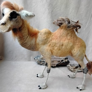 Op de afbeelding: Handgemaakte kameelsculptuur van bruine en beige wol. De kameel heeft een gedetailleerd gezicht met zwarte oogmarkeringen en een pluizige manen. De sculptuur staat op vier poten met gedetailleerde voeten.