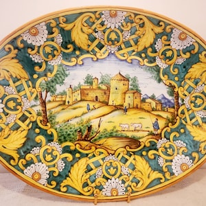 May include: An oval ceramic platter featuring a hand-painted scene of a small Italian village with a yellow, green and white floral border.