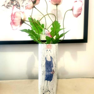 Può includere: Un vaso di ceramica bianca con un disegno dipinto a mano di una donna in un vestito blu. Il vaso è pieno di fiori rosa.