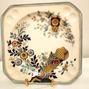 Puede incluir: Un plato cuadrado de cerámica para servir con un diseño floral azul y blanco. El plato tiene un borde elevado y un borde festoneado. El centro del plato presenta un diseño geométrico en tonos azules, amarillos y rojos.