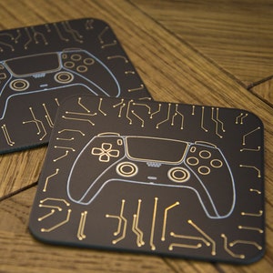 Puede incluir: Dos posavasos negros con un diseño de circuito impreso dorado y un contorno blanco de un mando de videojuego.