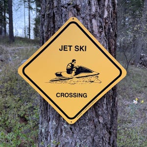 May include: A diamond-shaped yellow sign with a black border and black text that reads JET SKI CROSSING. The centre features a black graphic illustration of a person riding a jet ski, with small mounting holes at the top and bottom corners.