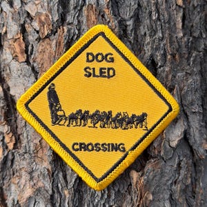 Pode incluir: Remendo bordado amarelo e preto com as palavras "DOG SLED CROSSING". O remendo apresenta uma silhueta preta de uma equipe de trenó puxado por cães e seu motorista. O remendo tem formato de losango e tem uma borda preta.