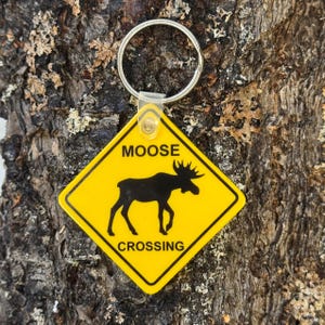 May include: Yellow and black moose crossing sign keychain. The keychain features a black silhouette of a moose on a yellow diamond-shaped sign. The words "MOOSE CROSSING" are printed on the sign. The keychain has a silver metal ring.