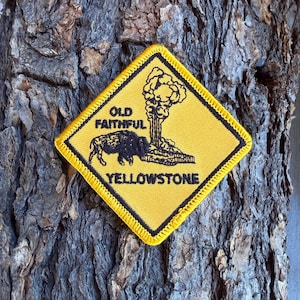 May include: A diamond-shaped yellow embroidered patch with a black border. The patch features a black graphic of a bison standing near a geyser eruption, with the text "OLD FAITHFUL" above the bison and "YELLOWSTONE" printed below the graphic.