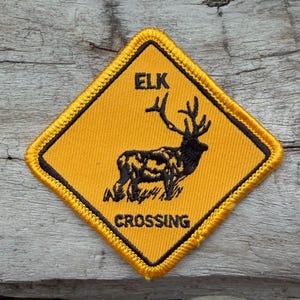 May include: A diamond-shaped embroidered patch with a yellow background and a black border. The patch features a black silhouette of an elk and the black text ELK CROSSING, mimicking the design of a standard road warning sign for wildlife.