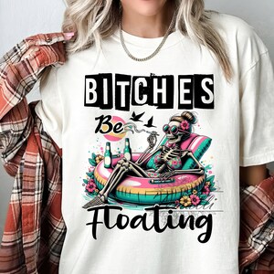 May include: White t-shirt with a graphic of a skeleton wearing sunglasses and a bikini, floating on a pink and green inflatable ring. The text "Bitches Be Floating" is printed on the shirt.