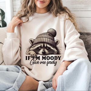 May include: A white sweatshirt with a graphic of a raccoon wearing a hat. The text on the sweatshirt reads "If I'm moody give me foody".