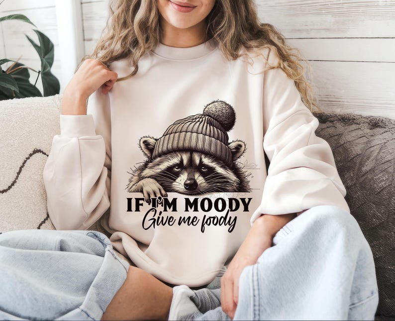 May include: A white sweatshirt with a graphic of a raccoon wearing a hat. The text on the sweatshirt reads "If I'm moody give me foody".