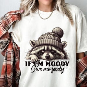 May include: White t-shirt with a graphic of a raccoon wearing a gray knit hat with a pom pom. The text "If I'm Moody Give me Food" is printed below the raccoon.