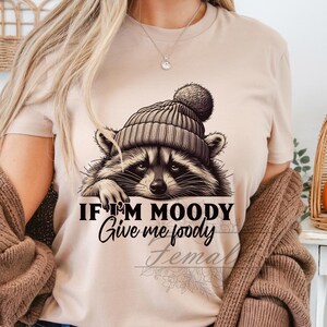 May include: A tan t-shirt with a raccoon wearing a brown and white hat. The text on the shirt reads "If I'm Moody Give me Foody".