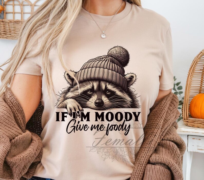 May include: A tan t-shirt with a raccoon wearing a brown and white hat. The text on the shirt reads "If I'm Moody Give me Foody".