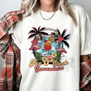 Puede incluir: Camiseta blanca con un diseño gráfico de una escena tropical con palmeras, flamencos y un cóctel. El texto "Five O'Clock Somewhere" está impreso en una fuente retro.