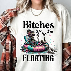 May include: White t-shirt with a graphic of a skeleton wearing a floral bikini and sunglasses, floating on a pink and blue inflatable ring. The text "Bitches Be Floating" is printed on the shirt.