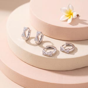 May include: Two pairs of silver-toned hoop earrings, each featuring a small bow-shaped charm encrusted with clear, sparkling stones. The earrings have a hinged closure mechanism and a polished metallic finish, presenting a refined and delicate appearance.
