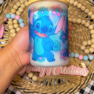 May include: A blue and white ceramic jar with a bamboo lid features a cartoon character with blue fur and large ears. The character is surrounded by pink and blue flowers. The jar is sitting on a woven surface.