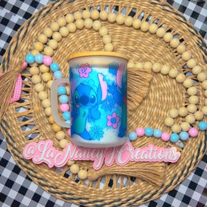 May include: A blue and white ceramic mug with a cartoon character design of Stitch from Lilo & Stitch. The mug has a wooden lid and a handle. The mug is sitting on a woven placemat with a pink tassel.