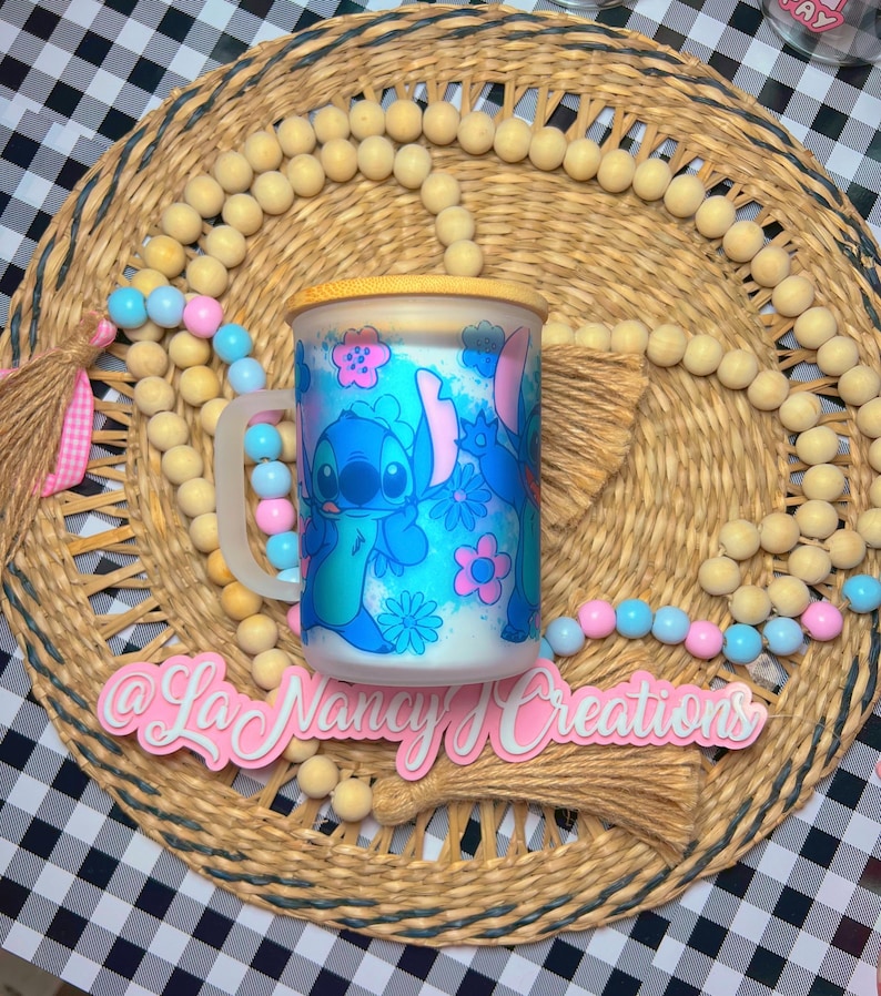 May include: A blue and white ceramic mug with a cartoon character design of Stitch from Lilo & Stitch. The mug has a wooden lid and a handle. The mug is sitting on a woven placemat with a pink tassel.