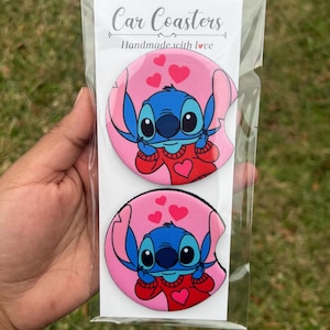 May include: Two pink car coasters with a blue Stitch character wearing a red sweater with hearts. The text "Car Coasters" and "Handmade with love" is printed on the packaging.