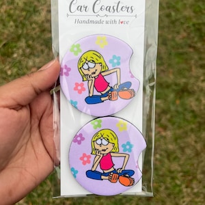 May include: Two purple car coasters with a cartoon girl design. The girl is wearing a pink shirt, blue jeans, and orange shoes. The coasters have a floral pattern with pink, yellow, green, and blue flowers. The text "Car Coasters" and "Handmade with love" is printed on the packaging.