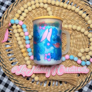 May include: A white ceramic mug with a blue and pink cartoon design of Stitch from Lilo & Stitch. The mug has a wooden lid and a handle. The mug is sitting on a woven mat with a pink tassel.