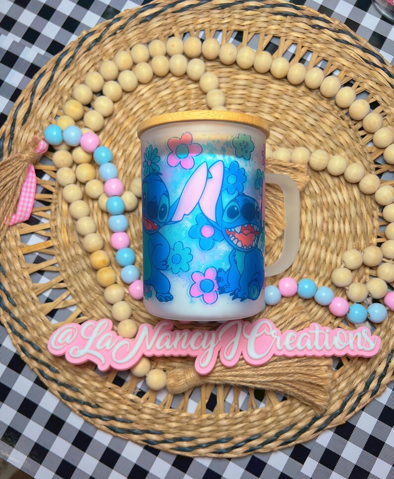 May include: A white ceramic mug with a blue and pink cartoon design of Stitch from Lilo & Stitch. The mug has a wooden lid and a handle. The mug is sitting on a woven mat with a pink tassel.
