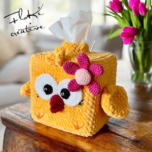 May include: A crocheted yellow chick tissue box cover with a pink flower on top. The chick has white eyes, a red beak, and white eyelashes. The tissue box cover is sitting on a wooden table.