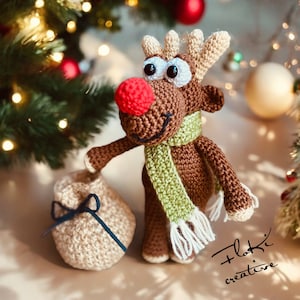 Könnte beinhalten: Ein braunes gehäkeltes Rentier mit einem grün-weißen Schal hält eine beige gehäkelte Tasche mit einem schwarzen Band. Das Rentier hat eine rote Nase und große braune Augen. Der Hintergrund ist ein Weihnachtsbaum mit roten Kugeln und Lichtern.